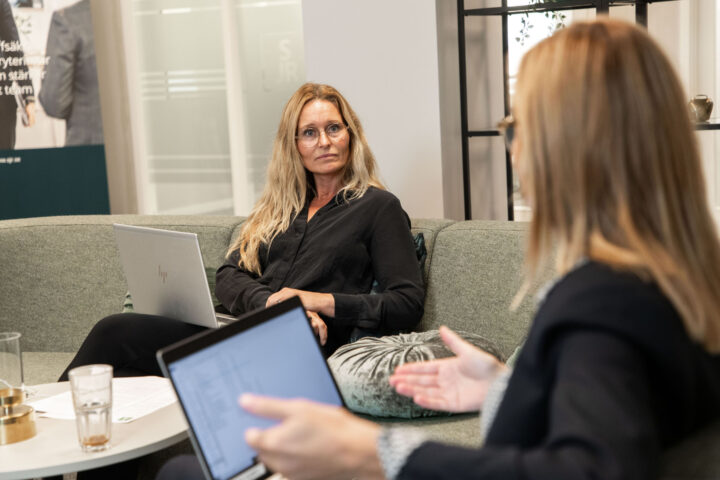 Två erfarna kvinnliga medarbetare från SJR har ett engagerat samtal vid sofforna i en loungemiljö med laptop och anteckningar, i ett ljust kontor i södra Sverige.