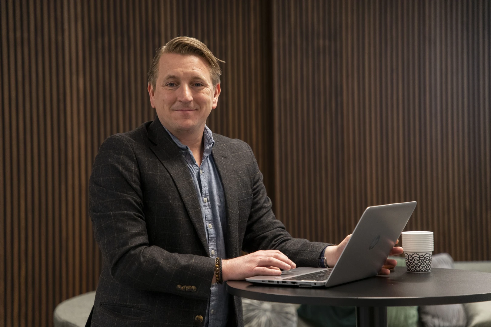En manlig medarbetare från SJR fokuserar på sitt arbete vid en stående arbetsstation med laptop och kaffekopp, i ett professionellt miljö med träpaneler i bakgrunden - Konsultpartner inom ekonomi & finans