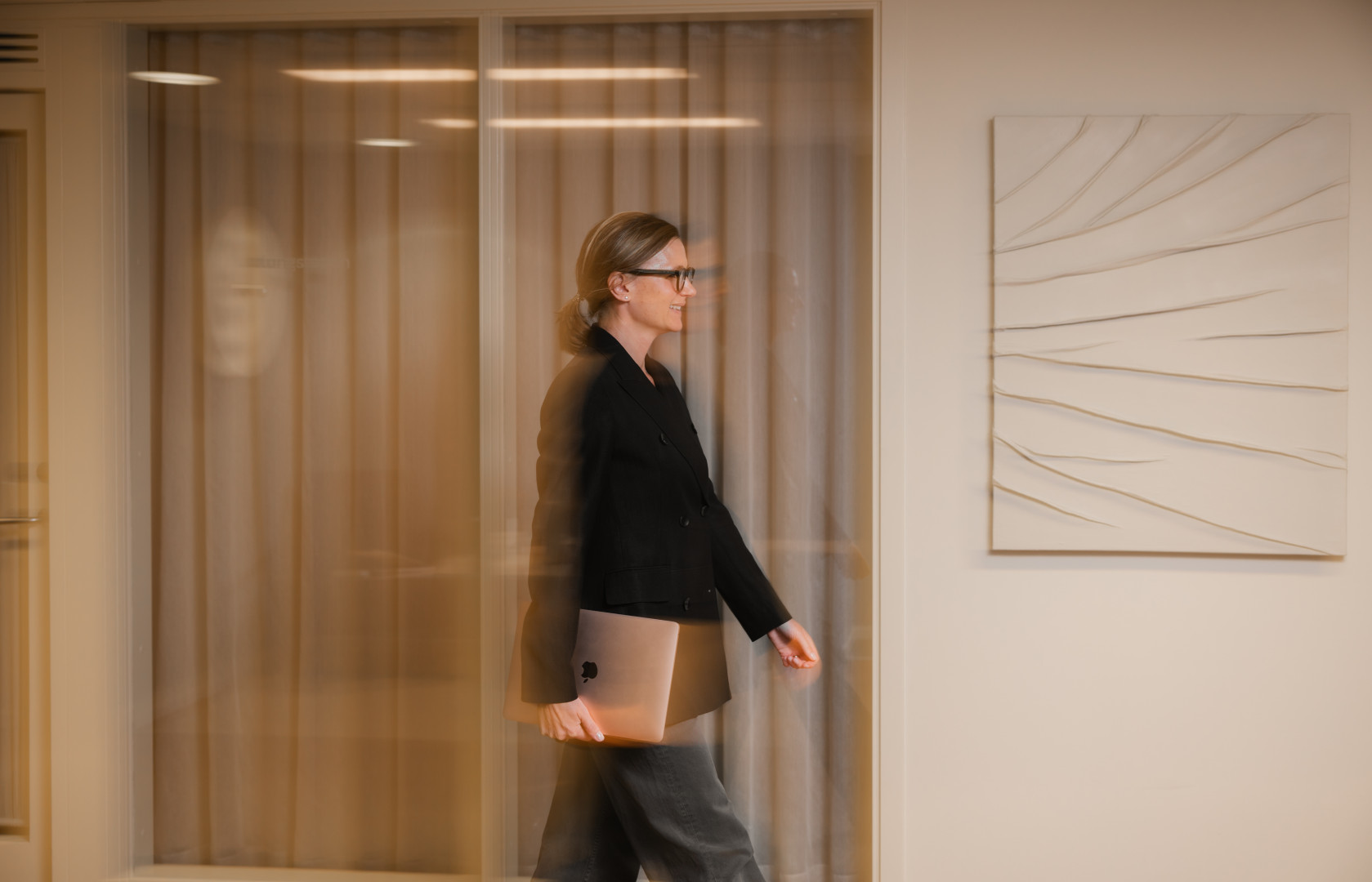 En medarbetare från SJR fångas i rörelse med en laptop i handen, på väg genom ett kontor med mjuka gardiner och modern konst i bakgrunden – en symbol för flexibilitet och framåtdriv.