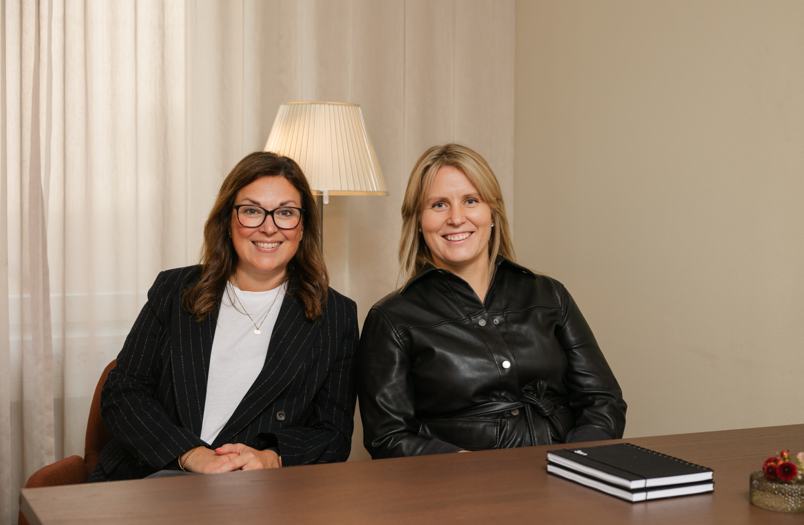 Rebecca och Elin, två representanter från sjr.se, sitter leende vid ett skrivbord i ett varmt inrett mötesrum med anteckningsbok och blomdekoration som förstärker den personliga och professionella atmosfären.