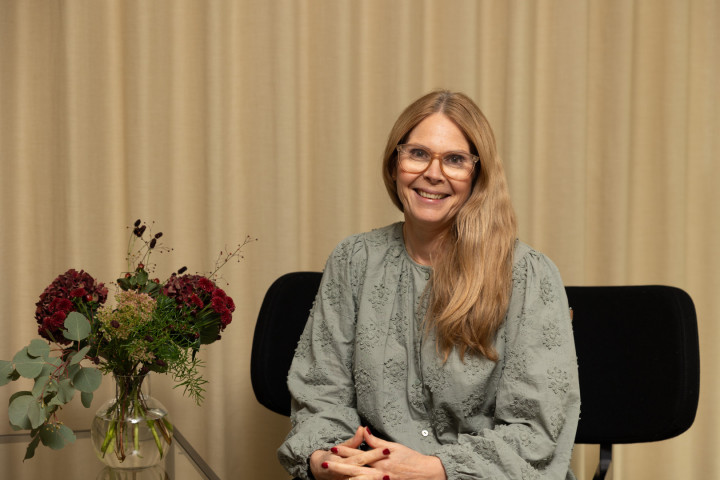 En kvinnlig medarbetare från SJR sitter leende i ett modernt kontor med gardiner i bakgrunden, vilket förstärker det personliga och tillgängliga uttrycket.