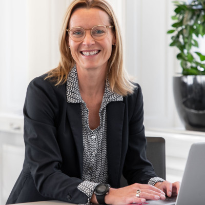 En leende kvinnlig kollega från SJR som sitter vid ett bord med sin laptop, klädd i kavaj och utstrålar professionalism och förtroendefulla relationer. Rekryteringsansvarig roll