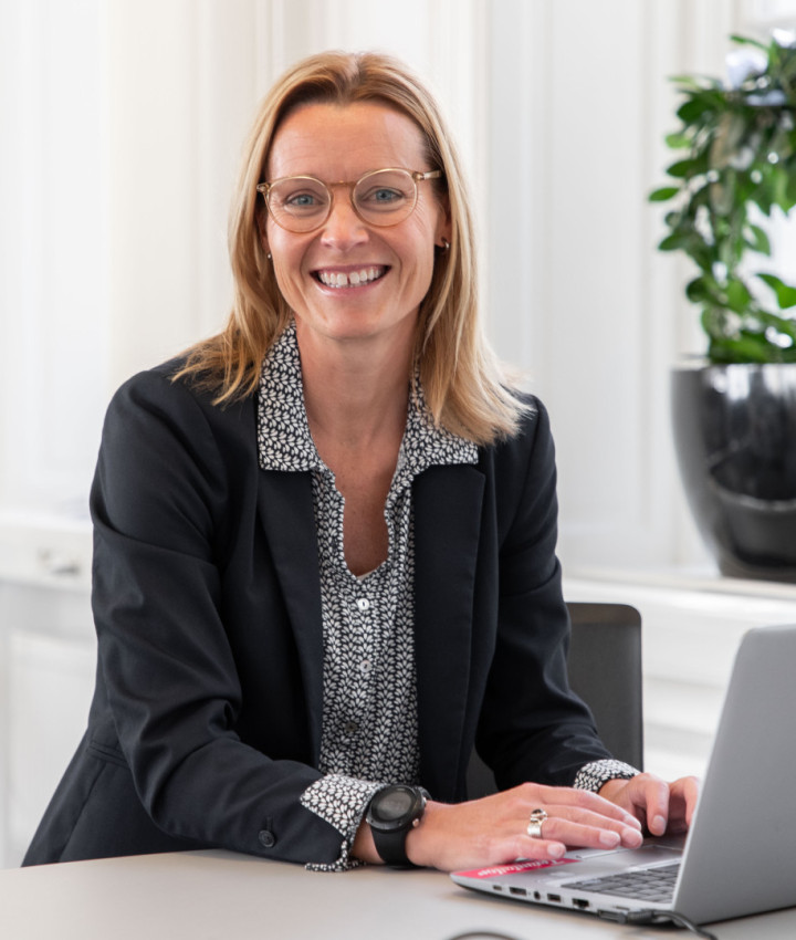 En leende kvinnlig kollega från SJR som sitter vid ett bord med sin laptop, klädd i kavaj och utstrålar professionalism och förtroendefulla relationer.