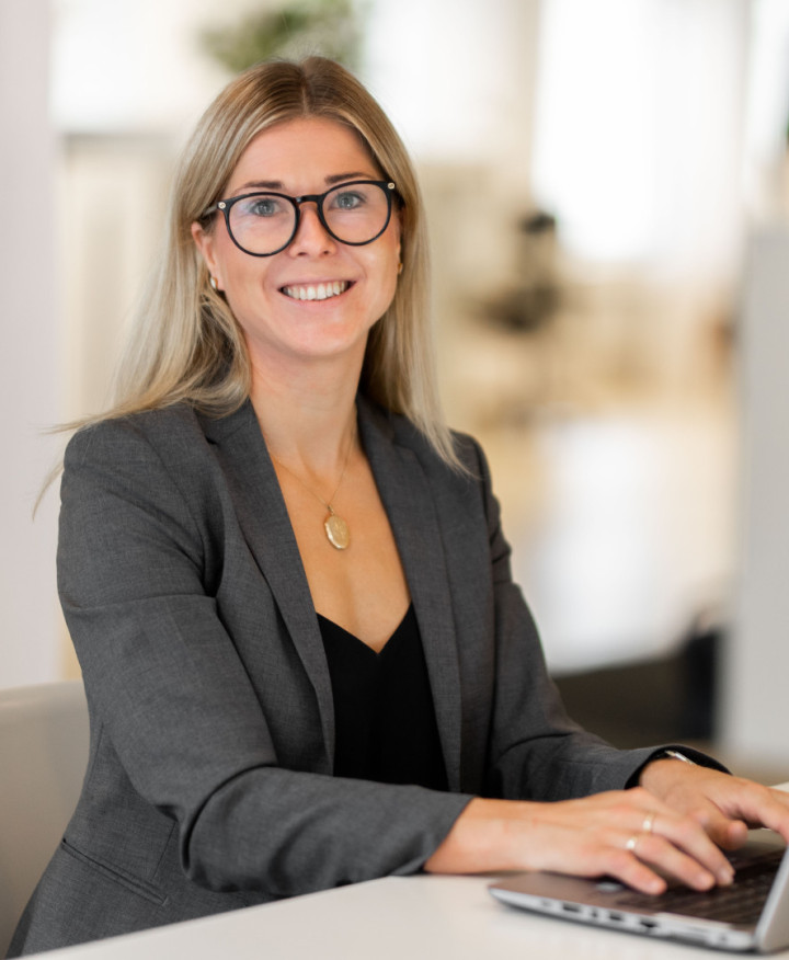 En leende kvinnlig kollega från SJR som sitter vid ett bord med sin laptop, klädd i kavaj och utstrålar professionalism och förtroendefulla relationer.